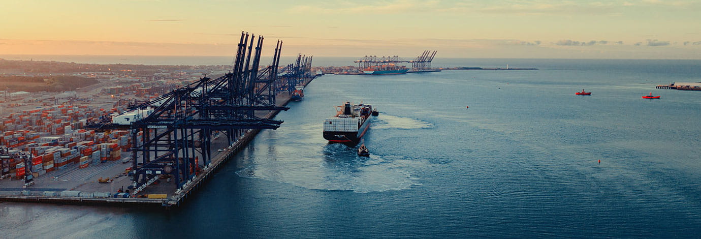 Sunrise over a container port, two tugs shepherd a container ship from under blue gantry cranes with rows of brightly colored shipping containers stacked behind them.